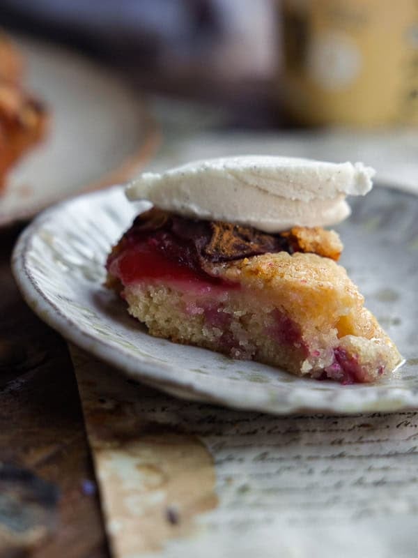 A slice of vegan plum torte with a golden crust and visible berries, topped with a scoop of vanilla ice cream, is served on a rustic ceramic plate.